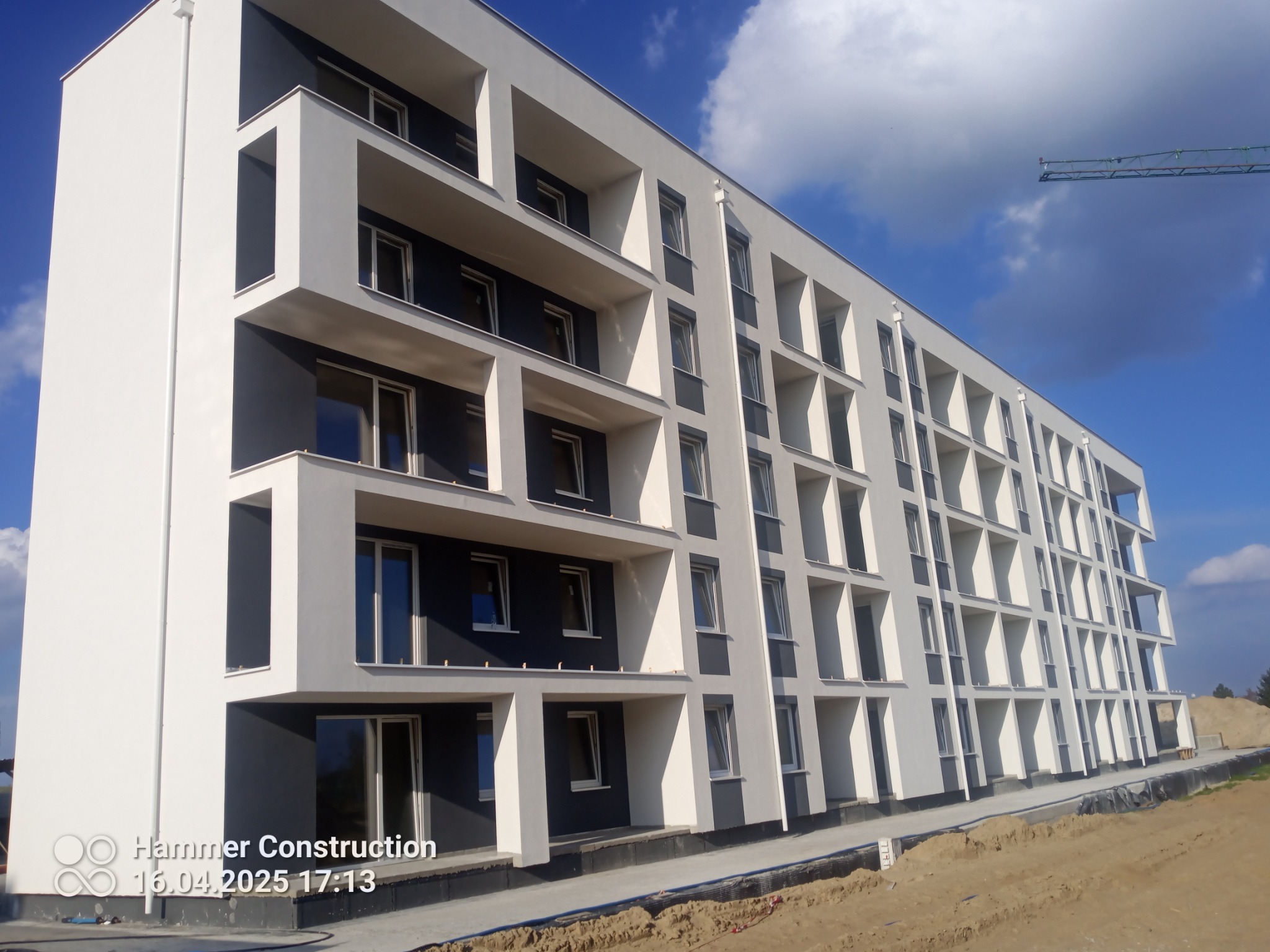 Nowoczesny, wielopiętrowy budynek z balkonami, pomalowany w odcieniach bieli i szarości, widziany z dołu pod kątem, z fragmentem żurawia budowlanego w tle.