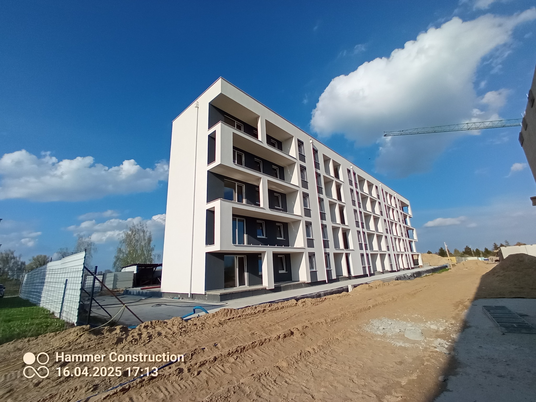 Nowoczesny, wielopiętrowy budynek z balkonami, świeżo po tynkowaniu elewacji, na tle błękitnego nieba z chmurami. Widoczny teren budowy i żuraw.
