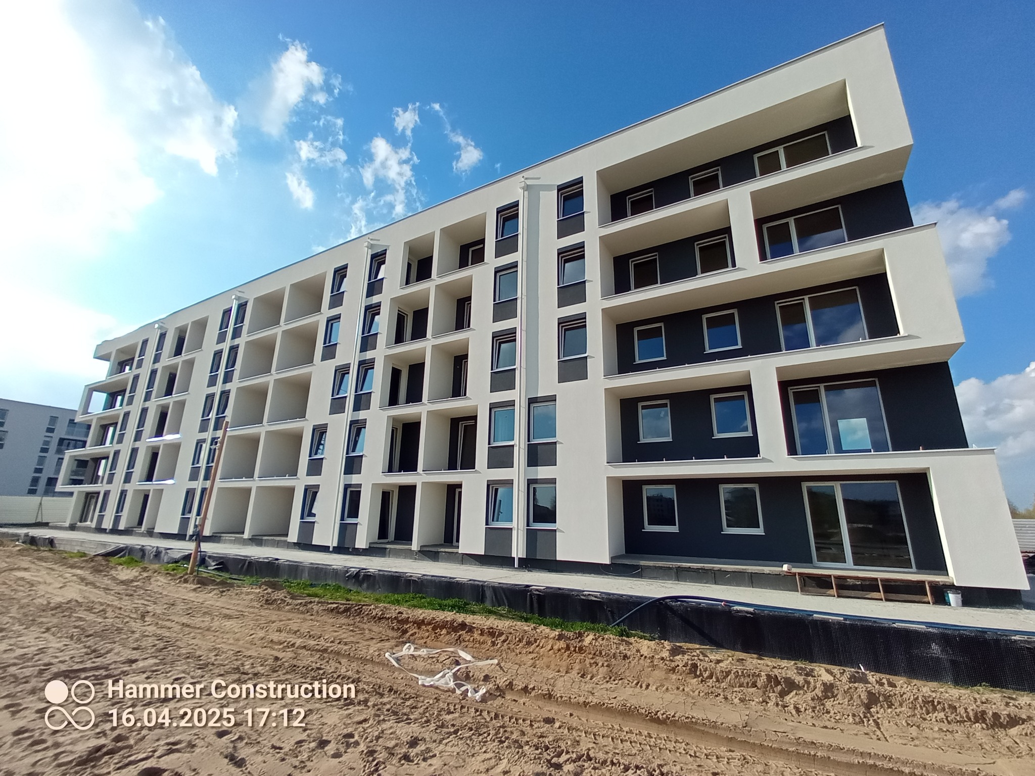 Nowoczesny, wielopiętrowy budynek z balkonami, świeżo po zakończeniu prac elewacyjnych, na tle błękitnego nieba. Widoczny teren budowy i logo firmy.