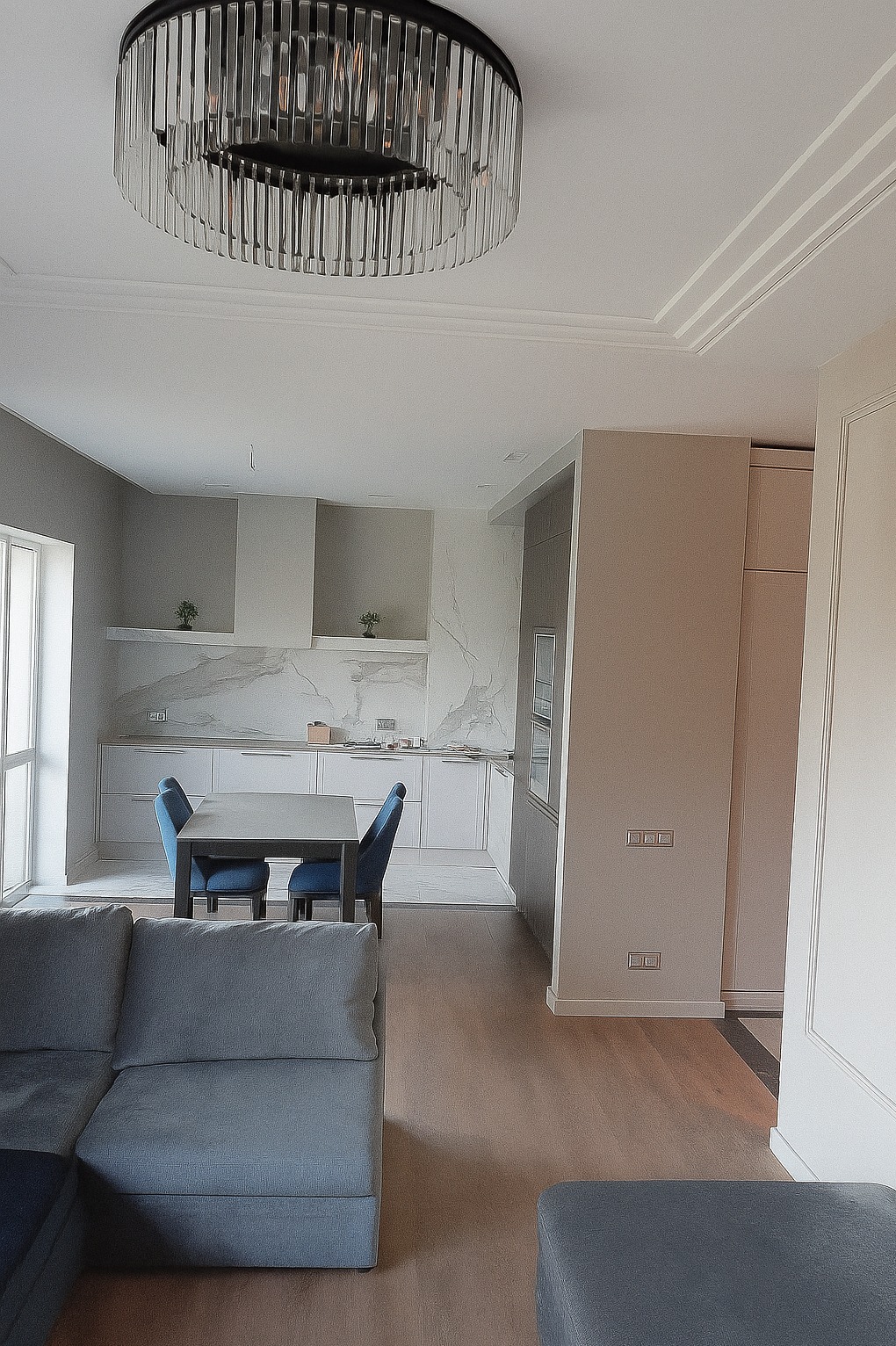 Nowoczesna kuchnia z marmurowym backsplashiem, oświetlona designerskim żyrandolem. Widoczny fragment salonu z szarą sofą. Jasne drewniane podłogi.