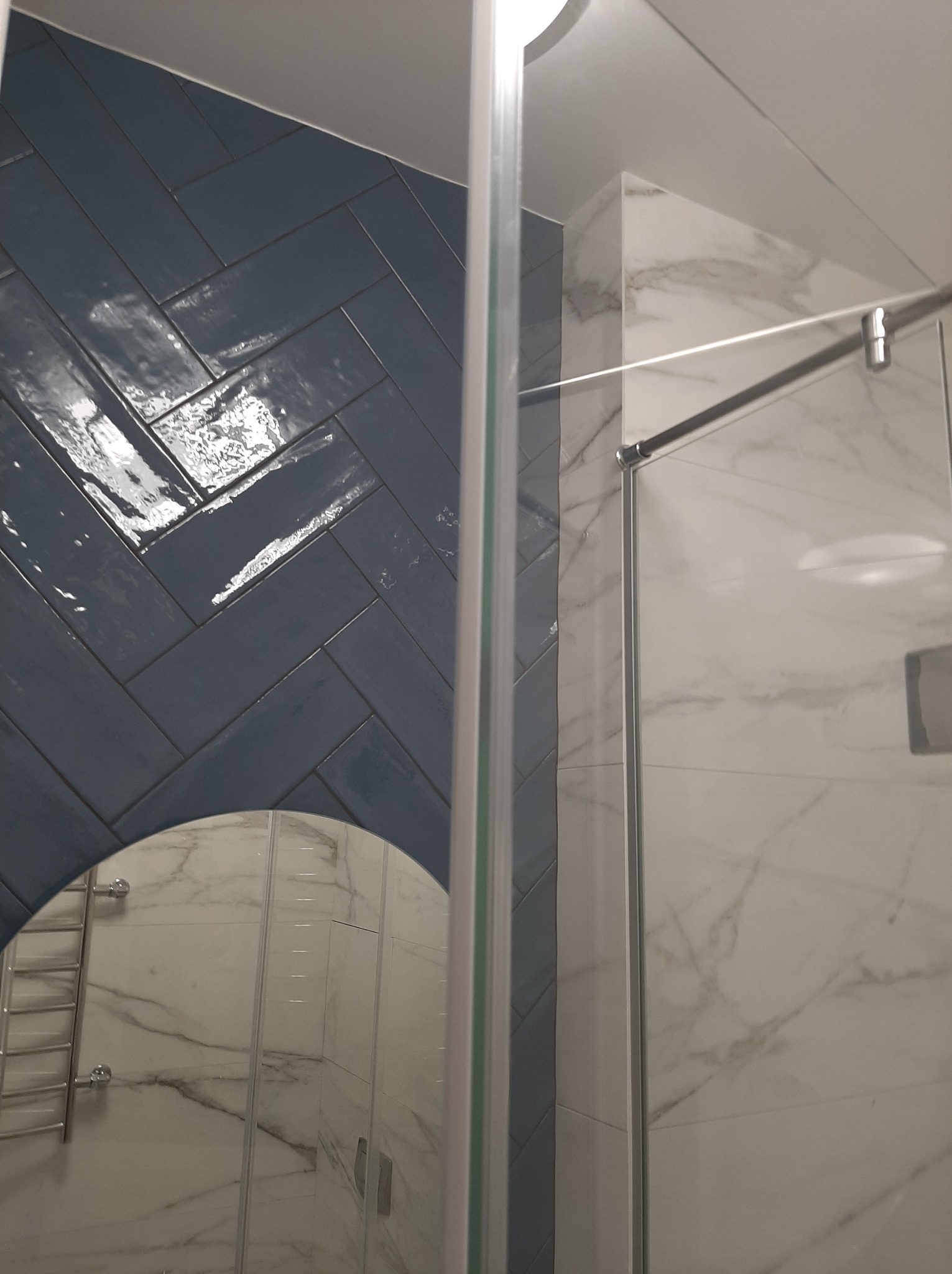 Błękitna ściana z płytek ułożonych w jodełkę w łazience, odbicie lustra i kabina prysznicowa z marmurowymi płytkami w tle.