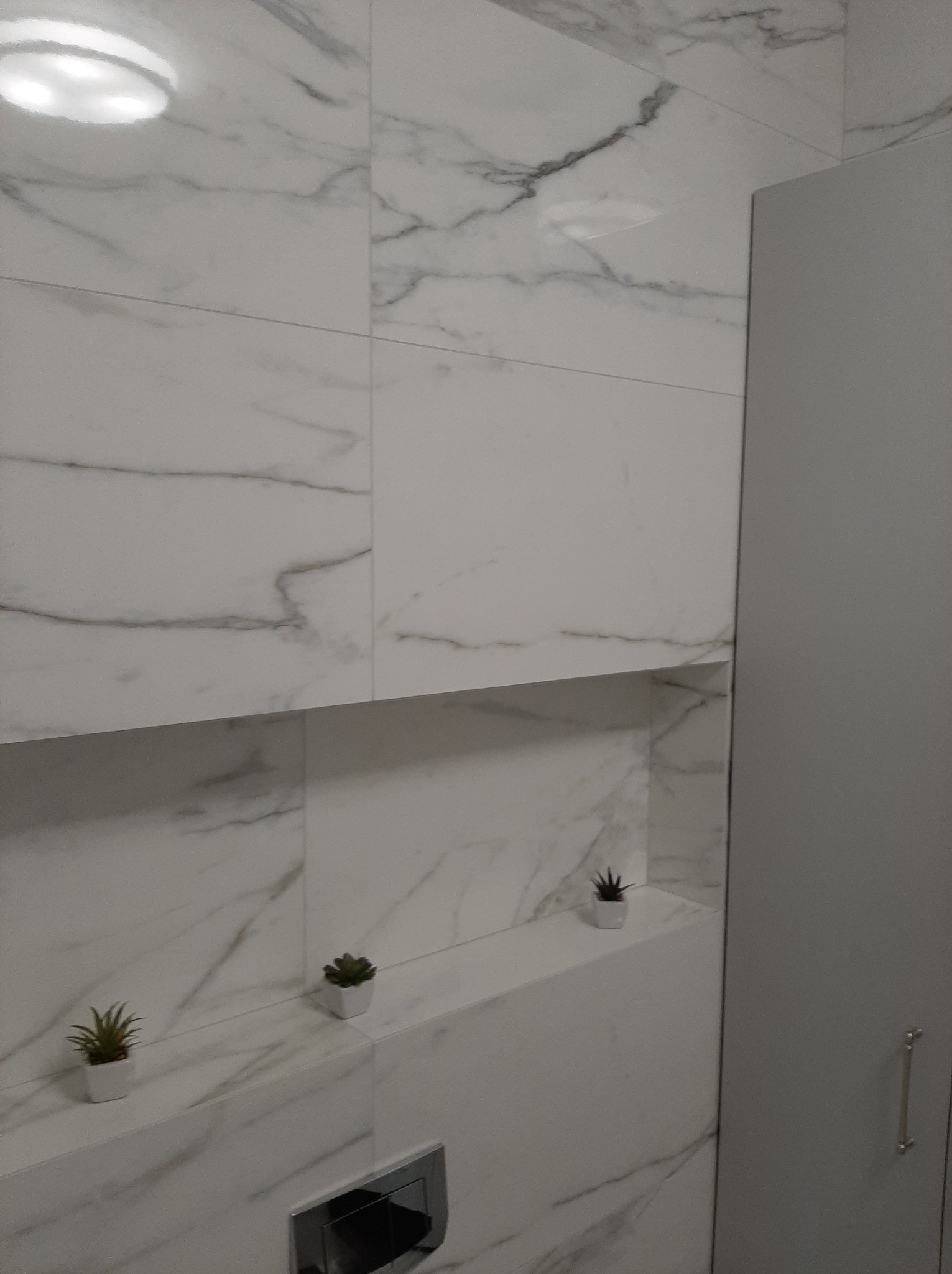 Biała łazienka z marmurowymi płytkami, wnęką z roślinami i szarą szafką. Minimalistyczny design i nowoczesne wykończenie.