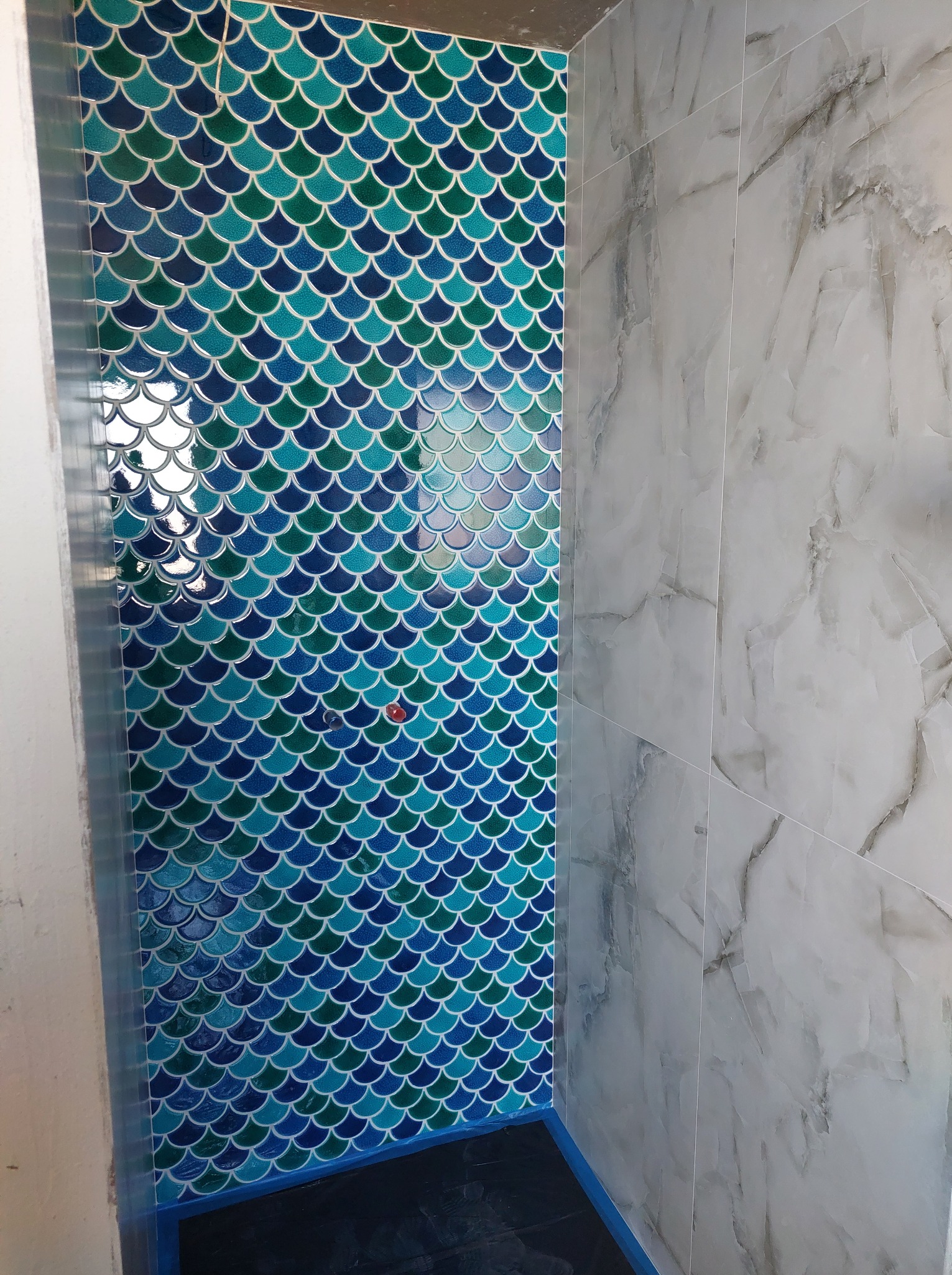 Ściana kabiny prysznicowej wyłożona mozaiką w kształcie rybiej łuski w odcieniach niebieskiego i zieleni, sąsiadująca z płytkami imitującymi marmur.