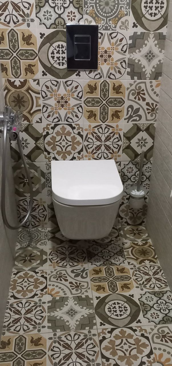 Wykończona łazienka z wiszącą toaletą i dekoracyjnymi płytkami w stylu patchwork na ścianach i podłodze. Czarny przycisk spłuczki i szczotka do WC.
