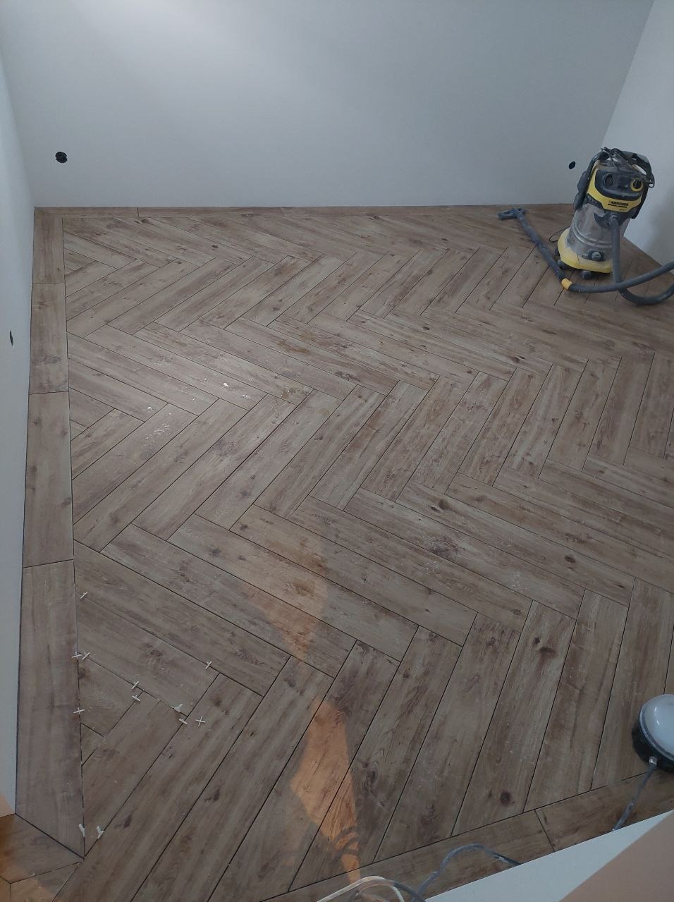 Ukończona podłoga z płytek imitujących drewno, ułożonych w jodełkę. Widoczny odkurzacz przemysłowy i krzyżyki dystansowe. Jasne ściany.