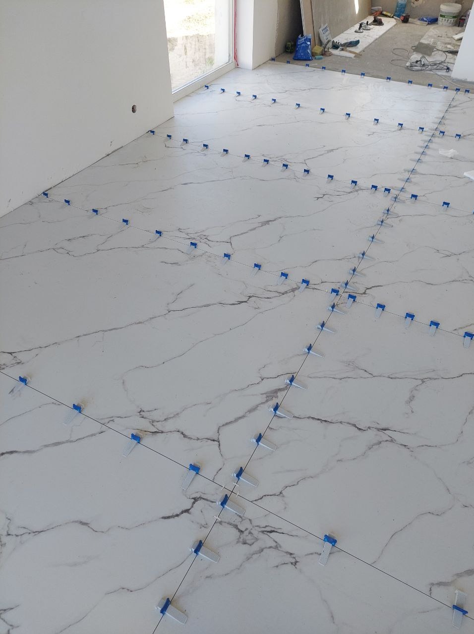 Układanie białych płytek imitujących marmur z niebieskimi klipsami poziomującymi na podłodze w trakcie remontu. Widok z góry, jasne wnętrze.
