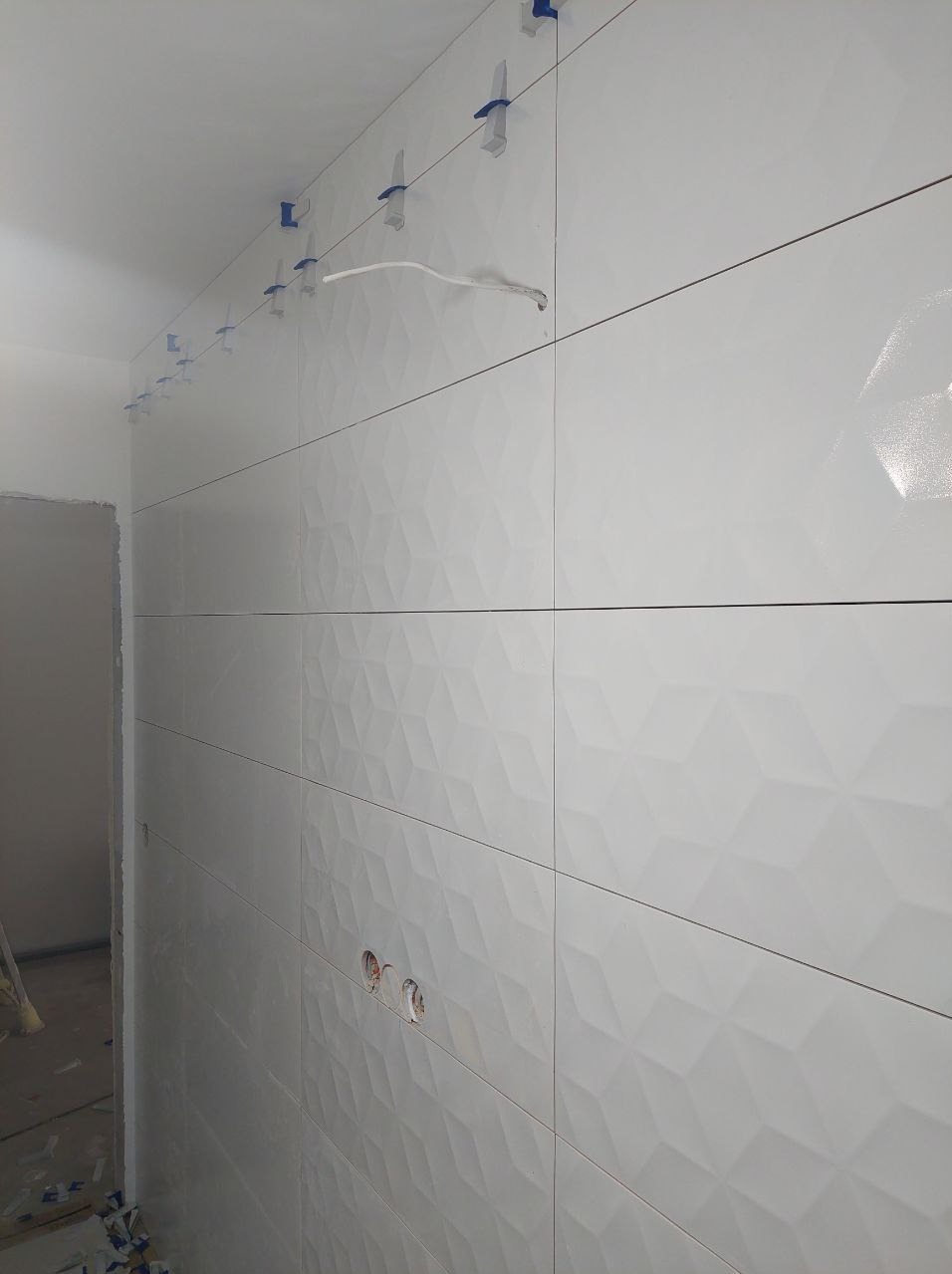 Białe, trójwymiarowe płytki ścienne w łazience, ułożone poziomo, z systemem poziomowania płytek. Widoczne otwory na gniazdka elektryczne i fragment sufitu.