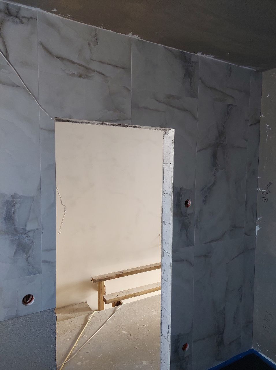Montaż płytek imitujących marmur w łazience, widoczny fragment ściany z otworem drzwiowym i instalacją elektryczną. Prace w toku, widoczne rusztowanie.