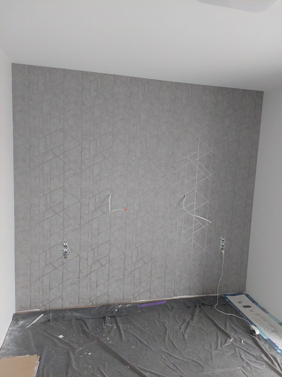 Ściana z tapetą w geometryczny wzór, widoczne gniazdka elektryczne i wystające przewody. Podłoga zabezpieczona folią malarską. Wnętrze w trakcie prac wykończeniowych.