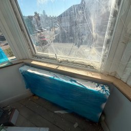 Janusz Kesicki - Okno w trakcie zabezpieczania folią malarską, z widocznym grzejnikiem osłoniętym folią. Przy podłodze leżą worki z klejem. Widok na zewnątrz.