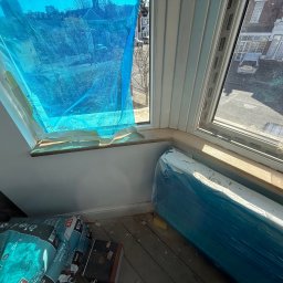 Janusz Kesicki - Okno zabezpieczone folią malarską, obok worki z klejem do płytek i grzejnik owinięty folią, przygotowanie do prac wykończeniowych w pomieszczeniu ze skosem.