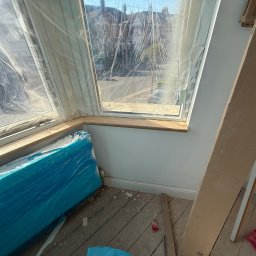 Janusz Kesicki - Nowe okno zabezpieczone folią, gotowe do prac wykończeniowych. Widoczna drewniana rama i skrzydła okienne. Podłoga drewniana, przygotowana do dalszych prac.