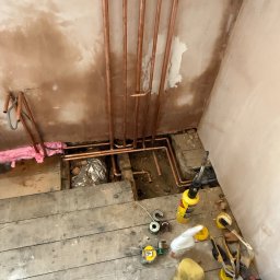 Janusz Kesicki - Miedziane rury grzewcze zamontowane przy ścianie z resztkami tynku, widoczne narzędzia instalacyjne na drewnianej podłodze. Instalacja CO w trakcie prac.