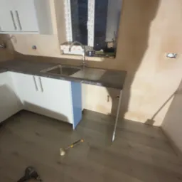 Montaż szafek kuchennych z szarym blatem i srebrnym zlewem. Widoczne narzędzia na podłodze, ściany przygotowane do malowania. Oświetlenie naturalne.