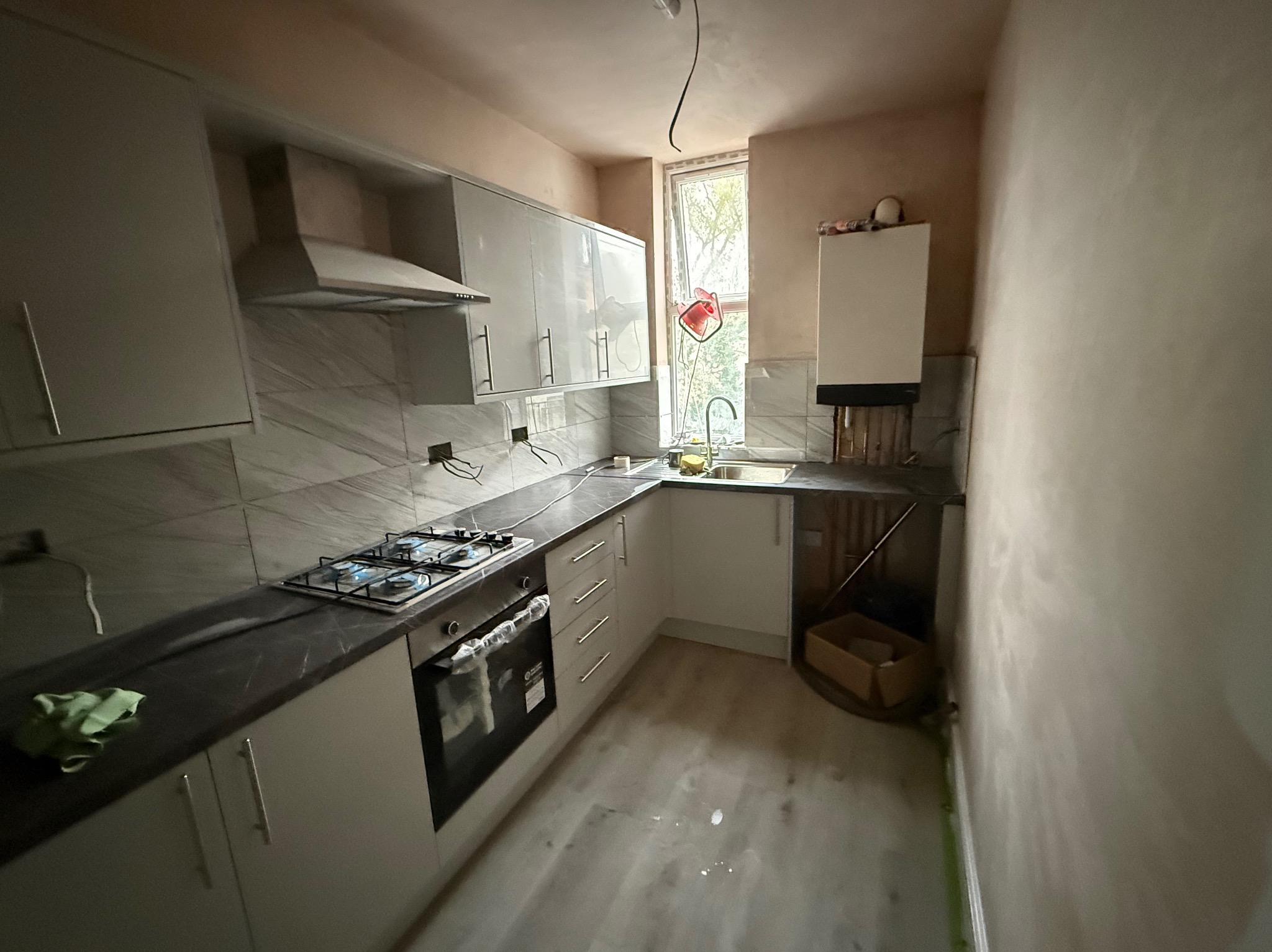 Jasna kuchnia w trakcie remontu: białe szafki, czarny blat, płyta gazowa. Ściany w gładzi, widoczne instalacje. Okno z widokiem na zieleń.