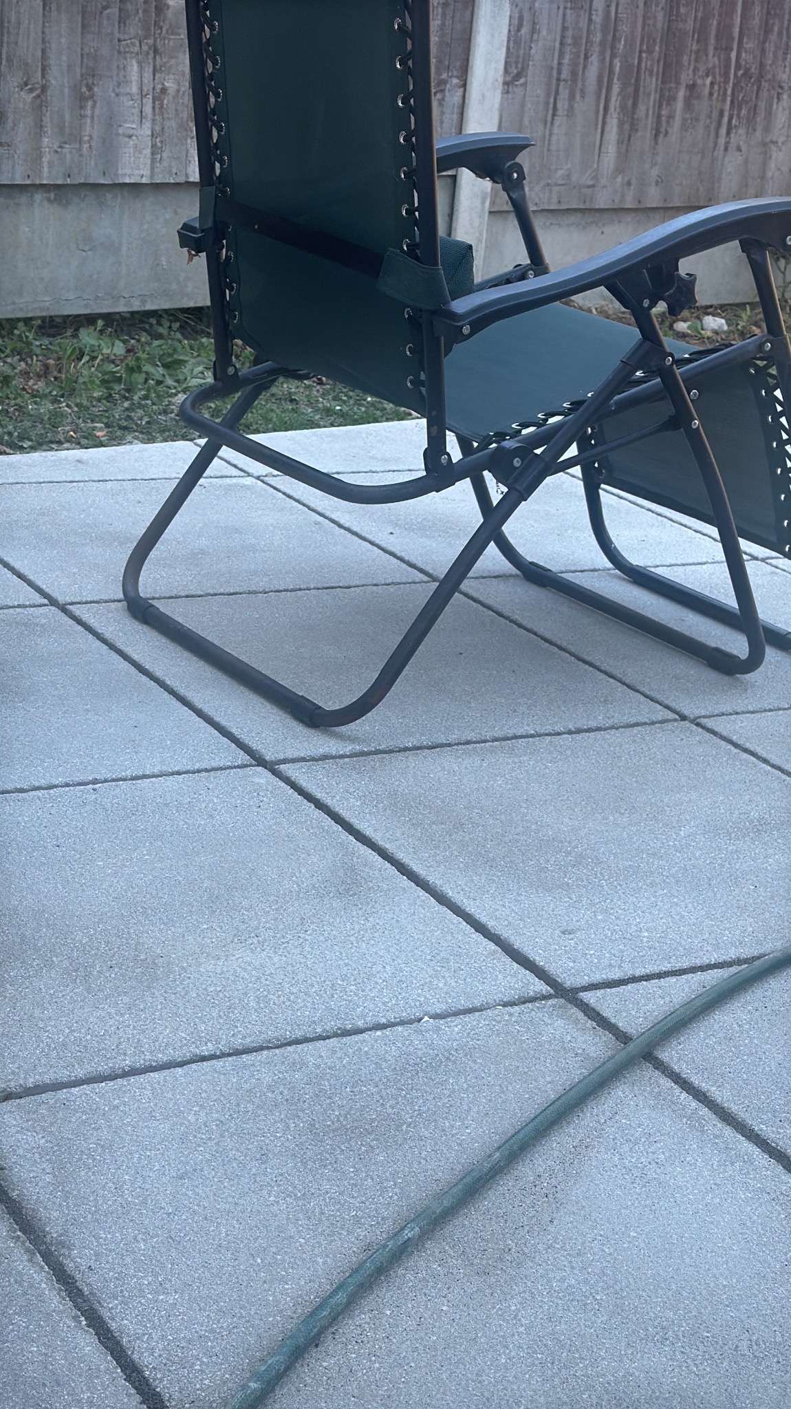 Ułożona szara kostka brukowa z zielonym krzesłem ogrodowym i fragmentem węża. Widoczne połączenia kostki i faktura materiału. Ujęcie z niskiego kąta.