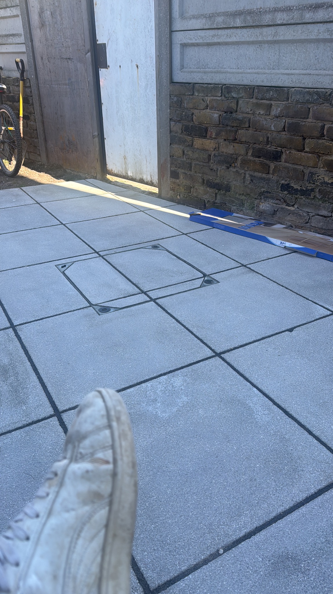 Nowa, szara kostka brukowa z włazem rewizyjnym w Ząbkowicach Śląskich. Widoczny fragment buta i narzędzia. Ułożona pod kątem, z ceglanym murem w tle.