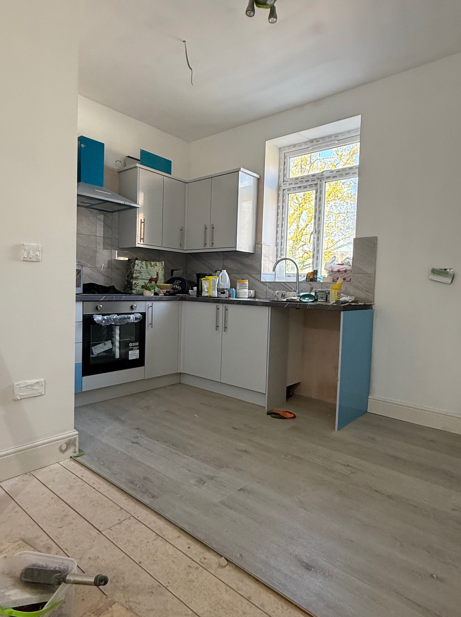Kuchnia w trakcie odświeżania: częściowo ułożone panele podłogowe, białe szafki z niebieskimi akcentami, okno z widokiem na zieleń. Widoczny etap prac.