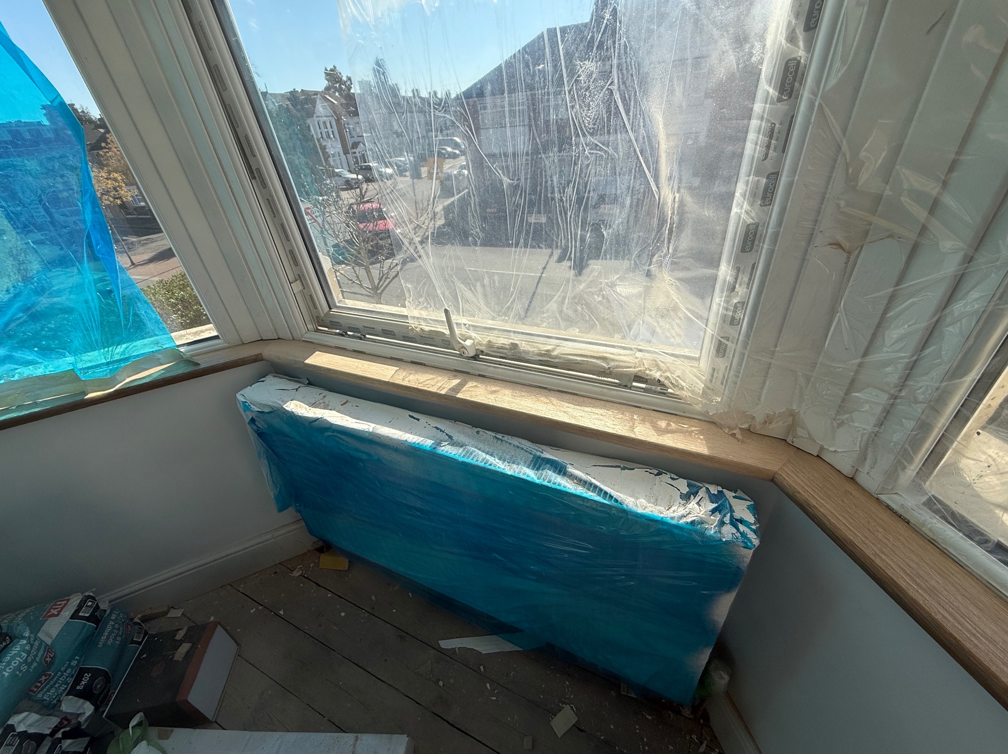 Okno w trakcie zabezpieczania folią malarską, z widocznym grzejnikiem osłoniętym folią. Przy podłodze leżą worki z klejem. Widok na zewnątrz.