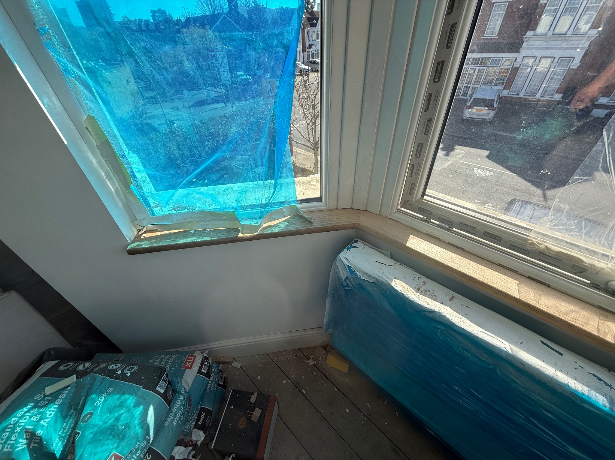 Okno zabezpieczone folią malarską, obok worki z klejem do płytek i grzejnik owinięty folią, przygotowanie do prac wykończeniowych w pomieszczeniu ze skosem.