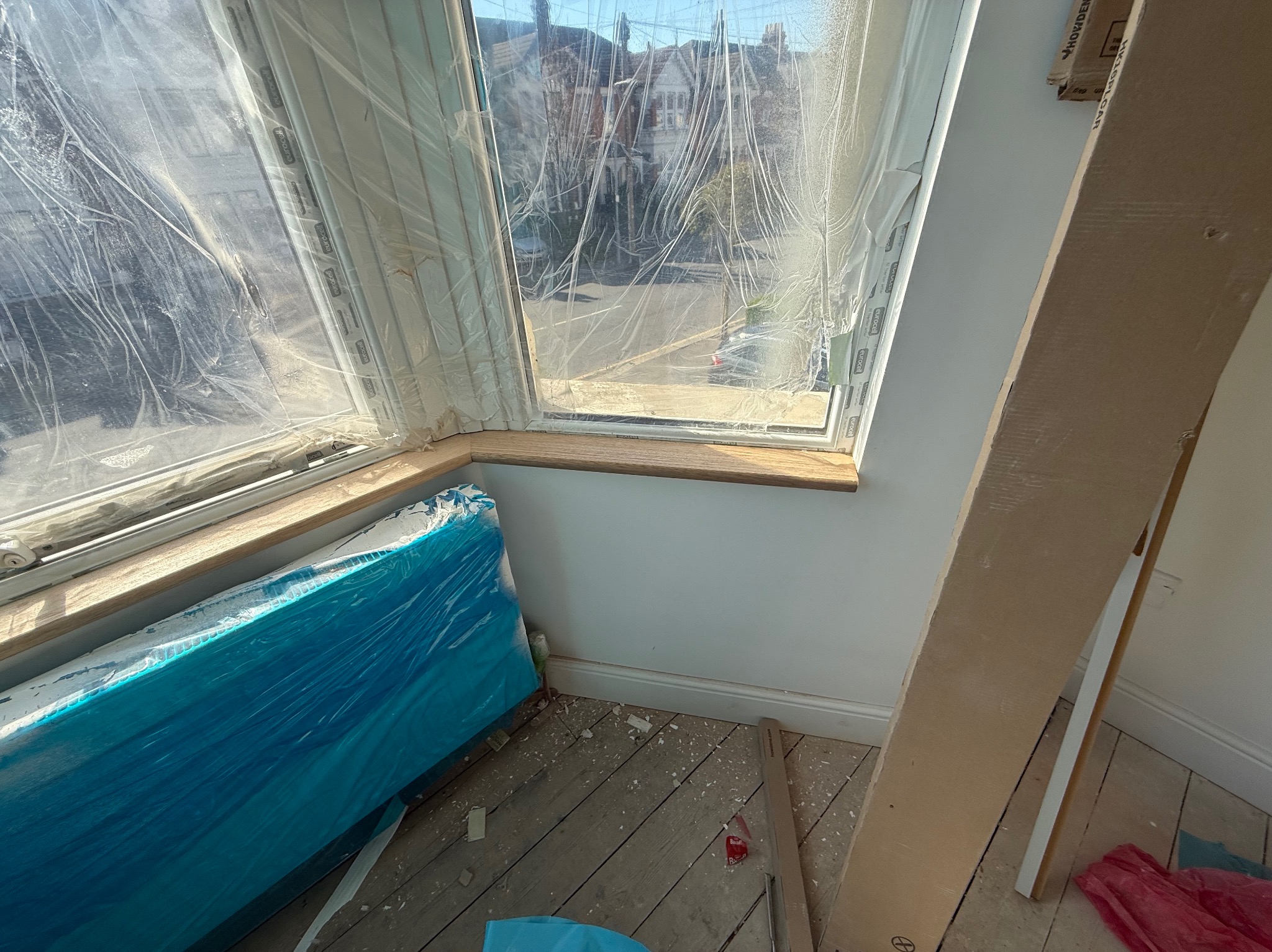 Nowe okno zabezpieczone folią, gotowe do prac wykończeniowych. Widoczna drewniana rama i skrzydła okienne. Podłoga drewniana, przygotowana do dalszych prac.