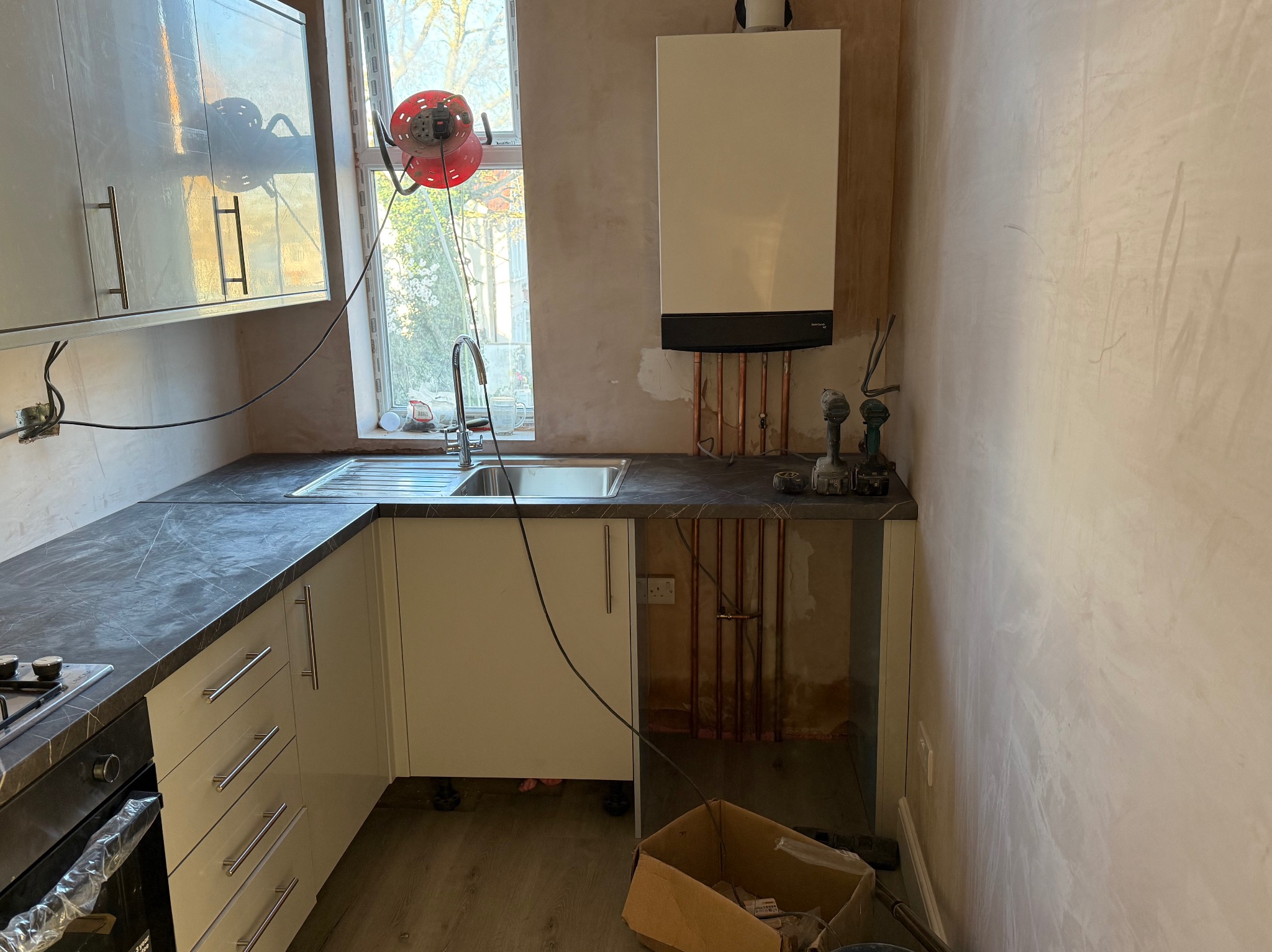 Wnętrze kuchni w trakcie prac: ściany po gładziowaniu, zamontowane szafki z blatem imitującym marmur, widoczny piec gazowy i narzędzia. Kabel zwinięty na czerwonym bębnie.