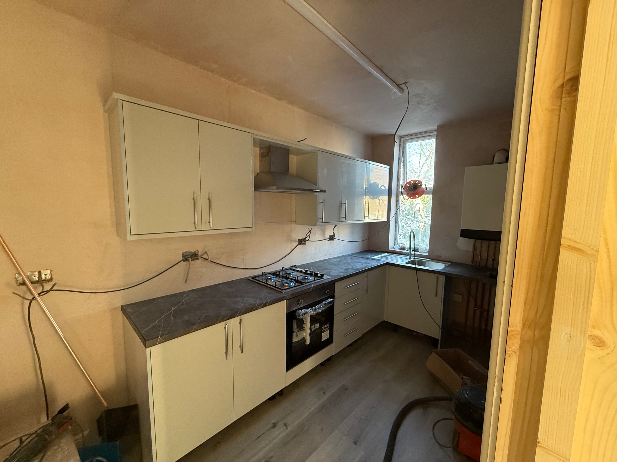 Jasna kuchnia w trakcie montażu: białe szafki z szarym blatem, płyta gazowa. Ściany w surowym tynku, widoczne kable, okno w tle. Wnętrze w trakcie wykończenia.