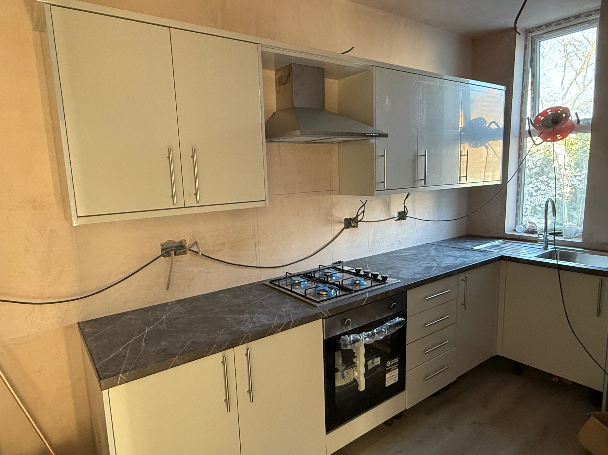 Kuchnia w trakcie montażu: białe szafki z szarym blatem, wbudowana płyta gazowa z niebieskim płomieniem, widoczne instalacje elektryczne, surowe ściany.