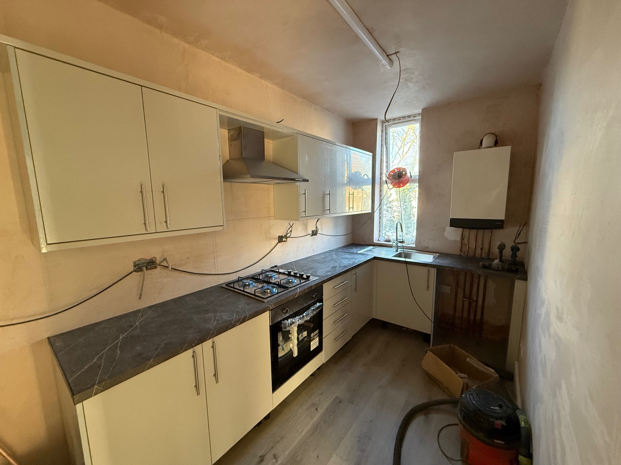 Jasna kuchnia w trakcie remontu: świeżo otynkowane ściany, częściowo zamontowane białe szafki z ciemnym blatem, widoczne instalacje. Podłoga wyłożona panelami.