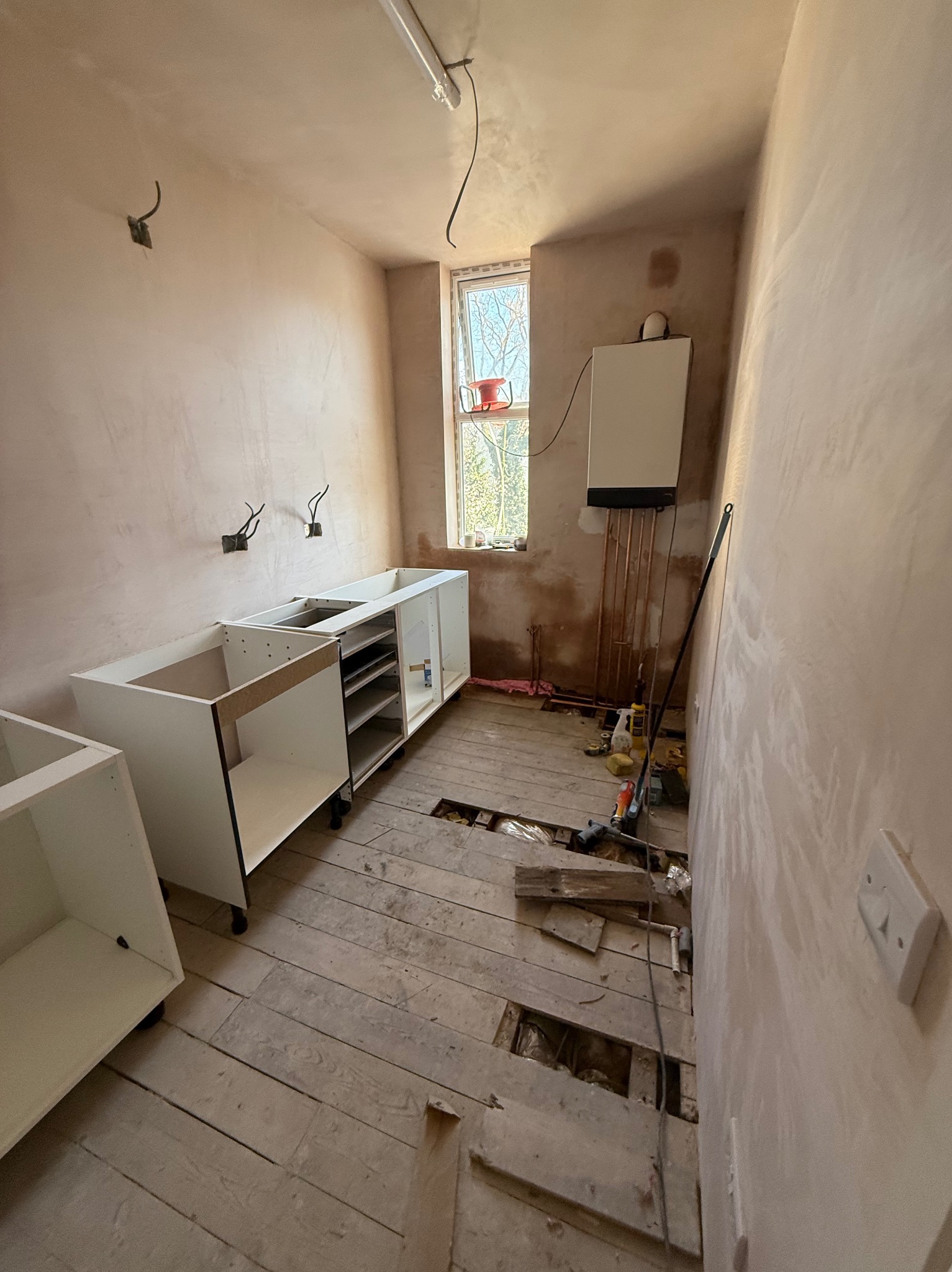 Wnętrze kuchni w trakcie remontu: ściany po gładzi, częściowo zamontowane białe szafki kuchenne, odsłonięta podłoga z desek, widoczny piec gazowy.