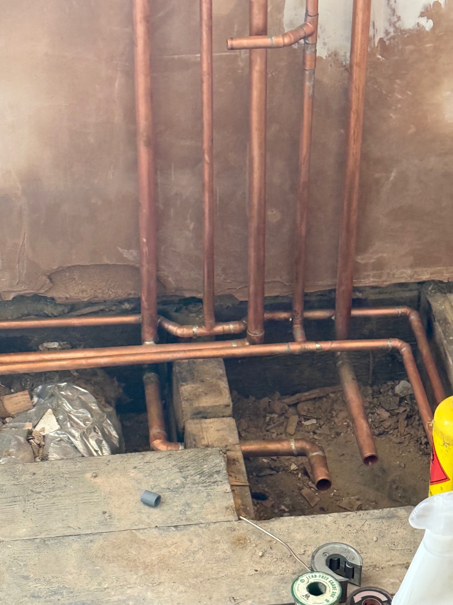 Instalacja miedziana: pionowe rury na tle surowej ściany, poziome rury w wykopie pod podłogą, narzędzia instalacyjne obok. Etap prac instalacyjnych.