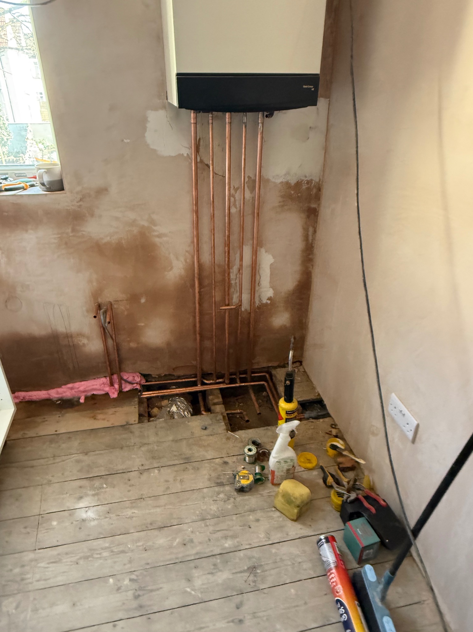Montaż pieca gazowego: miedziane rury łączą piec z instalacją, surowe ściany i podłoga z desek w trakcie remontu. Widoczne narzędzia i materiały.