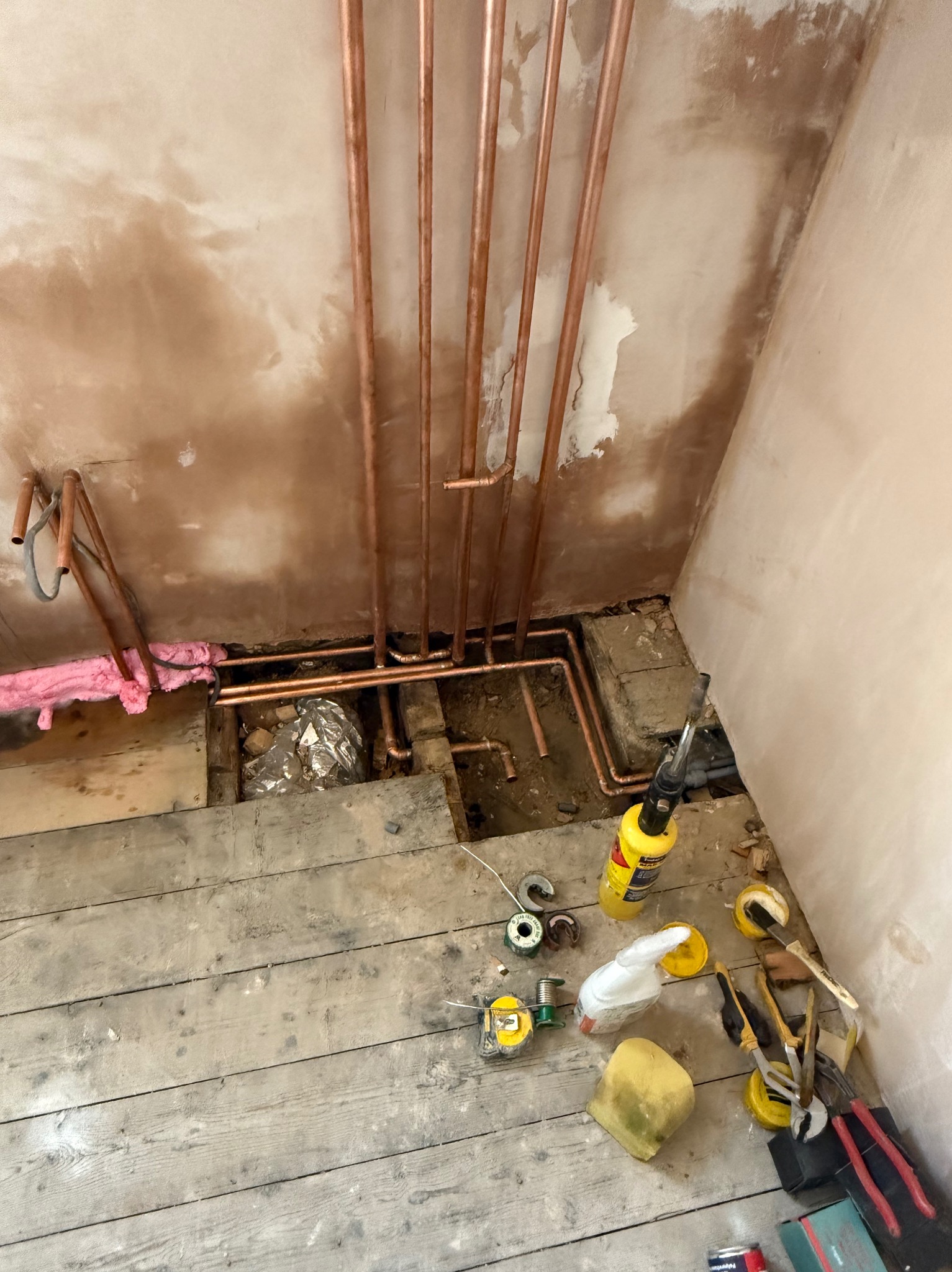 Miedziane rury grzewcze zamontowane przy ścianie z resztkami tynku, widoczne narzędzia instalacyjne na drewnianej podłodze. Instalacja CO w trakcie prac.