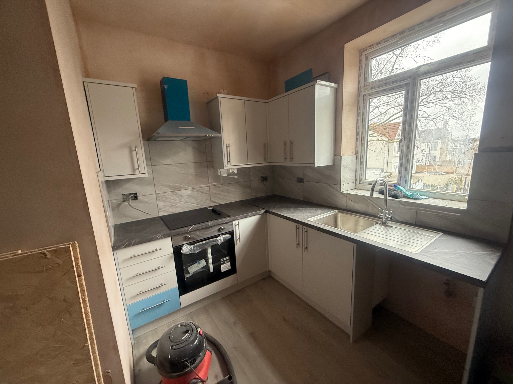 Jasna, narożna kuchnia z szarym blatem i płytkami. Dolne szafki z niebieskim akcentem, widoczne okno z widokiem na budynki. Ściany przygotowane do malowania.