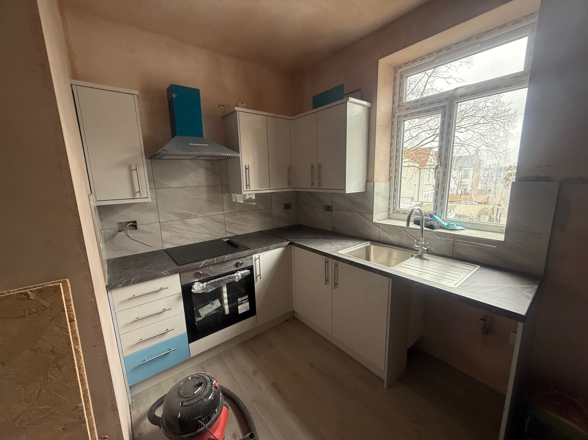 Jasna kuchnia z niebieskimi akcentami w trakcie wykańczania: białe szafki, szary blat, okno z widokiem na drzewa. Ściany przygotowane pod malowanie.