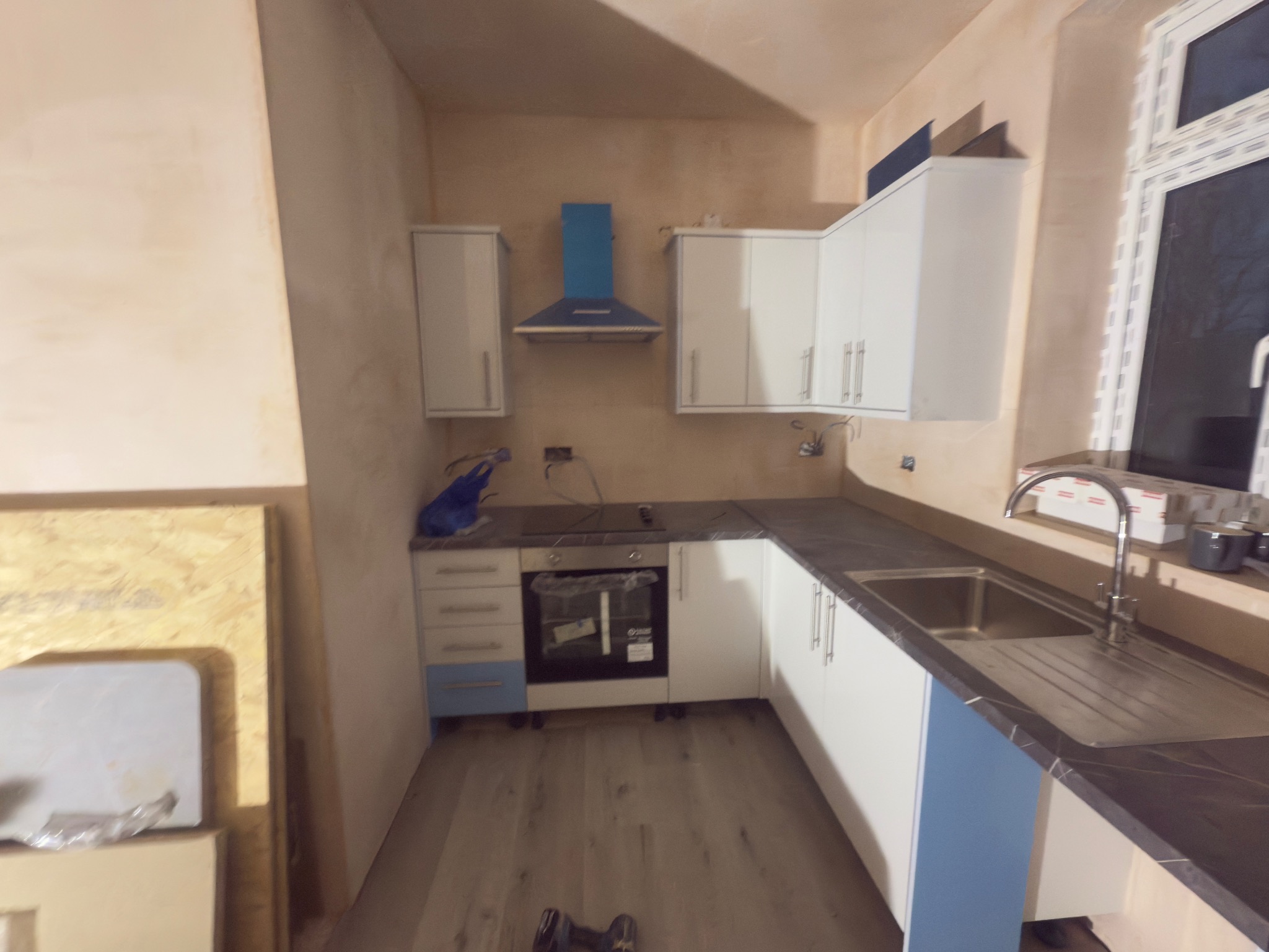 Wnętrze kuchni w trakcie montażu: białe szafki z niebieskimi akcentami, okap, zlew z baterią i fragment podłogi z panelami. Ściany w stanie surowym.