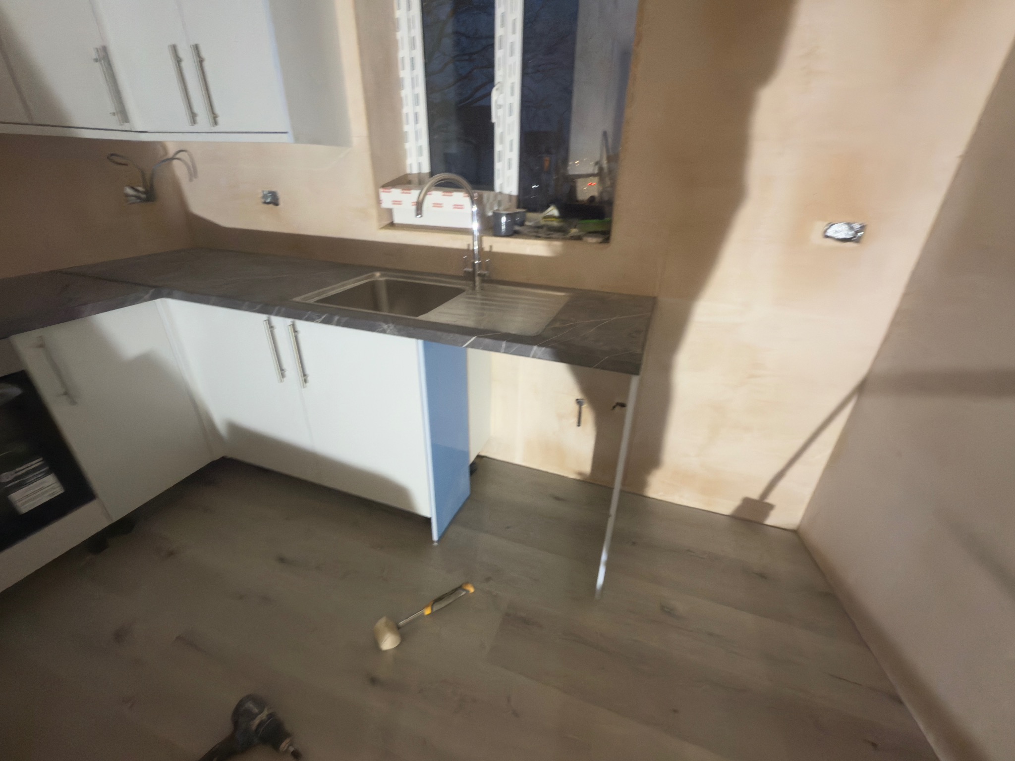 Montaż szafek kuchennych z szarym blatem i srebrnym zlewem. Widoczne narzędzia na podłodze, ściany przygotowane do malowania. Oświetlenie naturalne.