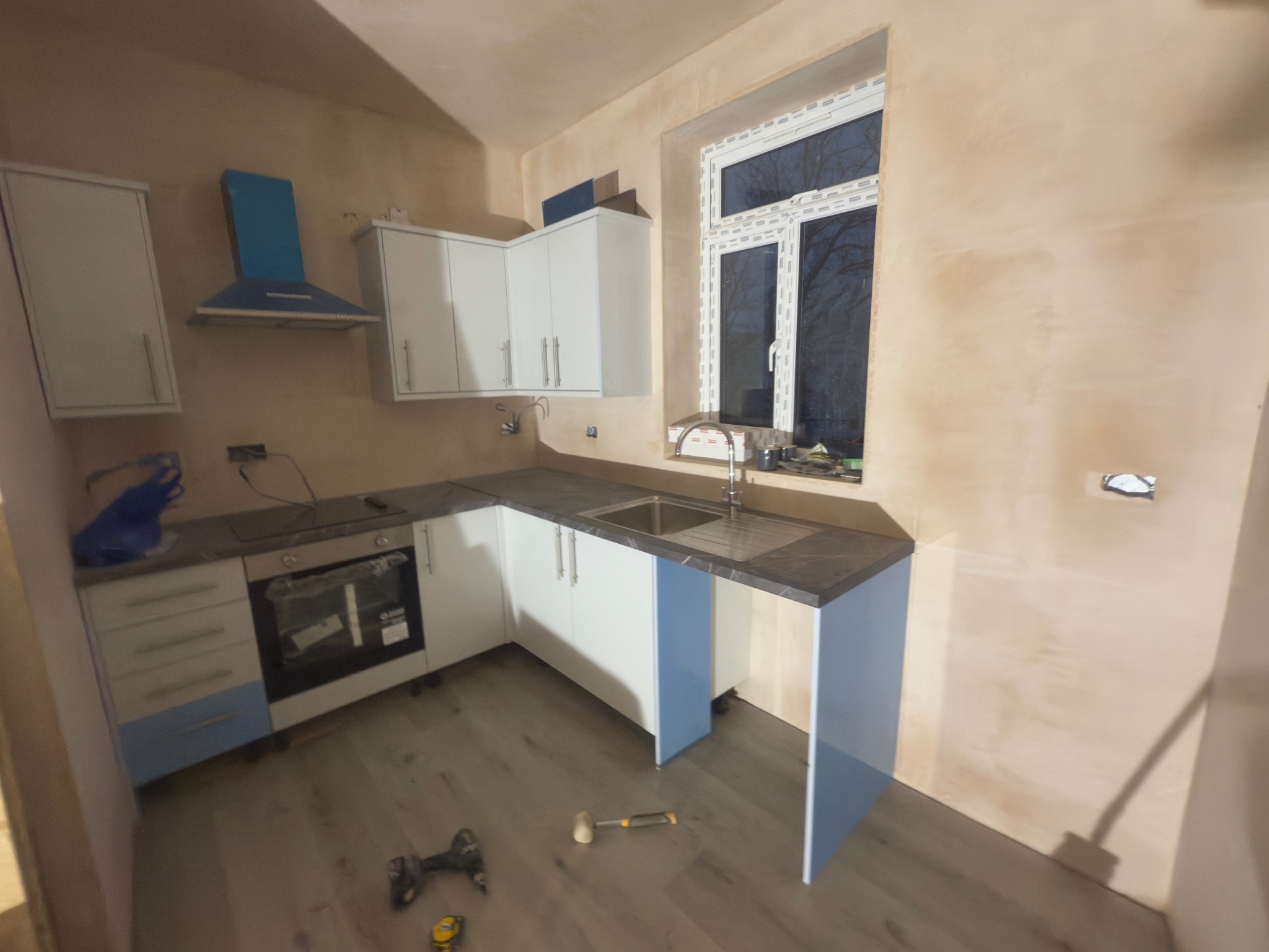 Wnętrze kuchni w trakcie remontu: białe szafki, szary blat, okno. Ściany w surowym tynku. Na podłodze leżą narzędzia.