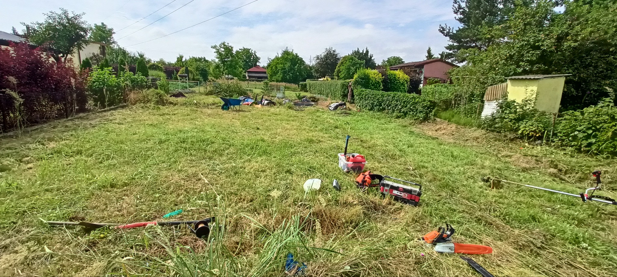 Rozległy teren z częściowo skoszoną trawą, widoczne narzędzia ogrodnicze: kosiarka, piła łańcuchowa i podkaszarka. W tle zabudowania i zieleń.