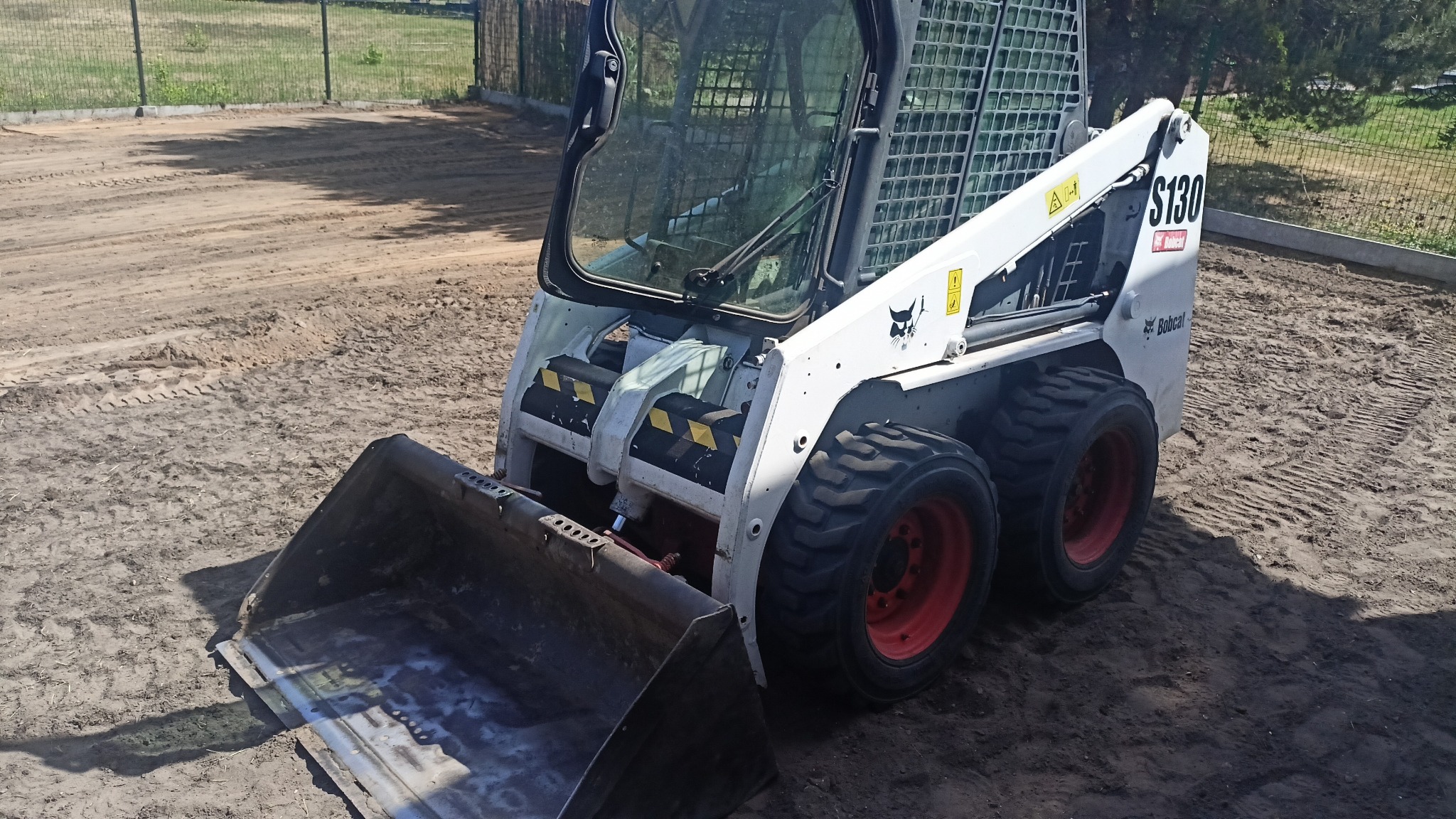 Kompaktowa ładowarka Bobcat S130 na piaszczystym podłożu. Widoczne ślady opon i świeżo wyrównany teren, sugerujące prace ziemne. Sprzęt gotowy do dalszych działań.
