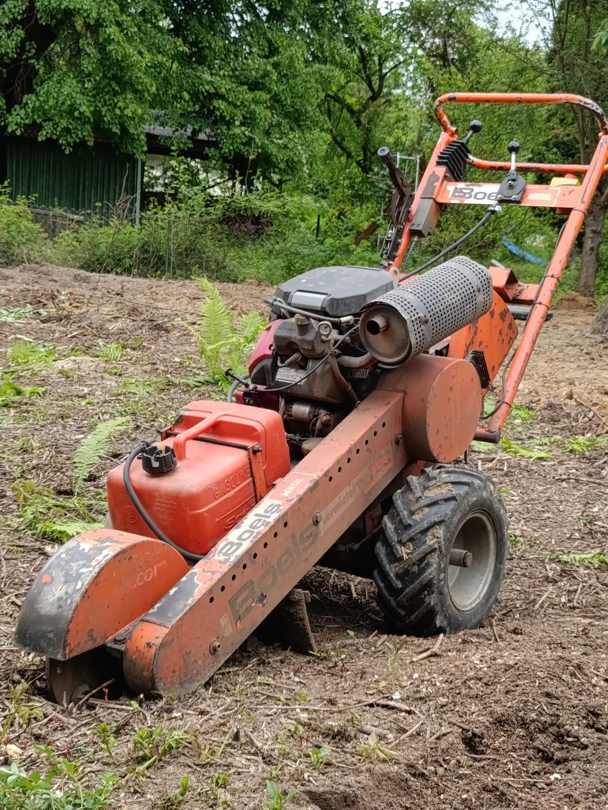 Freza do pni Boels na terenie po wycince, widoczne resztki gałęzi i pniaków. Maszyna z silnikiem spalinowym, pomalowana na pomarańczowo, w tle zieleń.