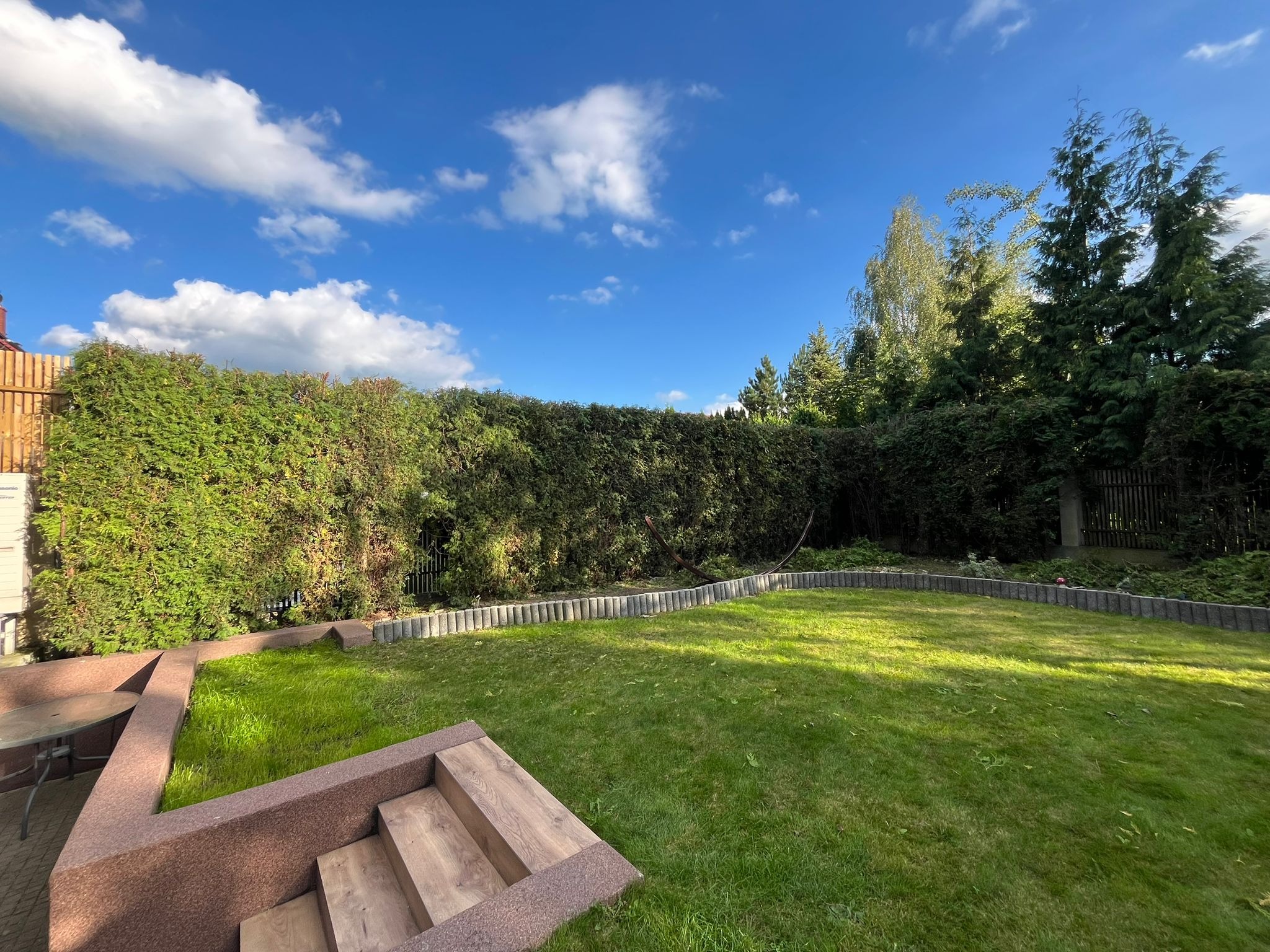 Zadbany trawnik z równo przyciętym żywopłotem w słoneczny dzień. Widoczne elementy architektury ogrodowej: schody i obrzeża trawnika.