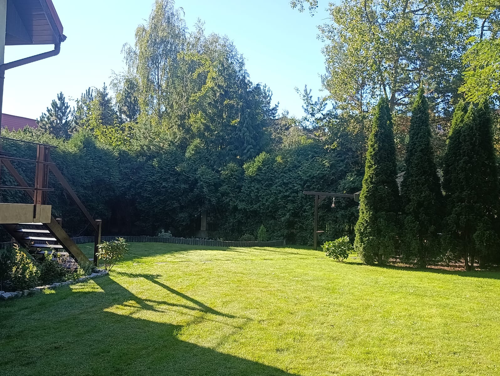 Zadbany trawnik z równo przystrzyżoną trawą, otoczony zielenią drzew i krzewów. Widoczne schody tarasowe i elementy dekoracyjne ogrodu. Słoneczny dzień.