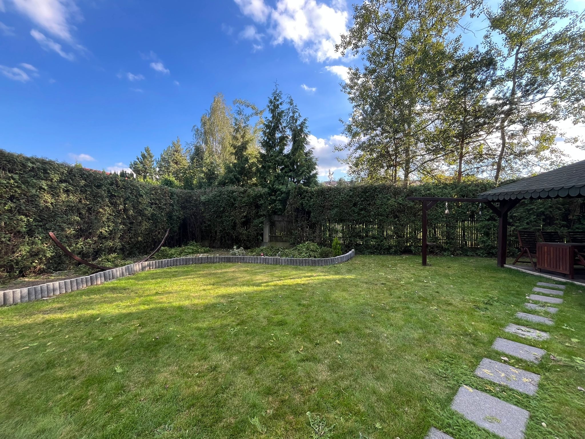 Zadbany trawnik z kostką obrzeżną i ścieżką z płyt. W tle żywopłot, drzewa i altana. Słoneczny dzień, błękitne niebo z chmurami.