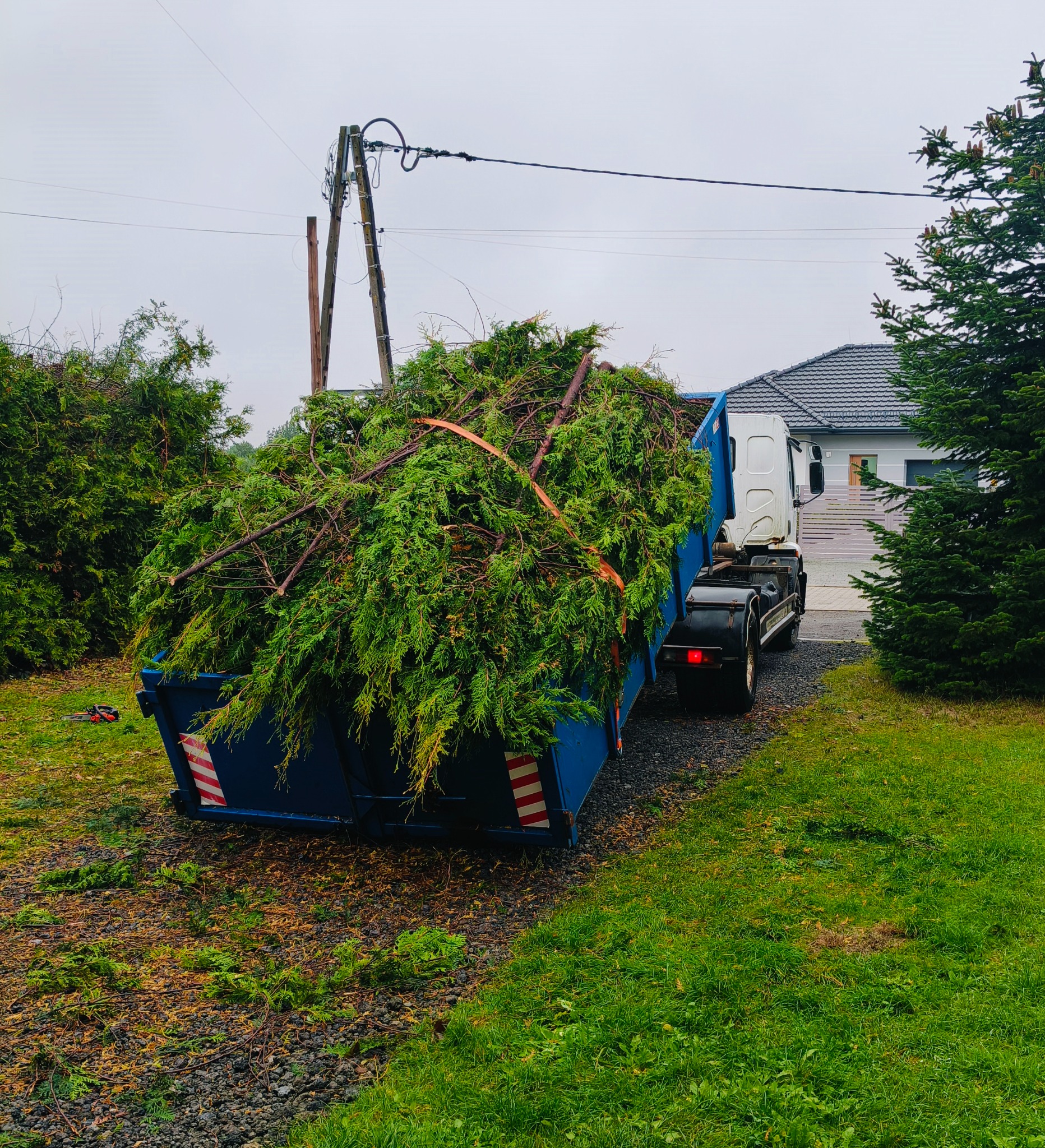 Kontener wypełniony gałęziami i ściętymi drzewami, stojący na posesji przed domem. Widoczny fragment ciężarówki i drzewa iglaste.