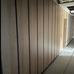 MAR-BUD - Wykończona ściana z paneli drewnopodobnych z czarnymi wstawkami, widok na sufit z instalacjami w surowym stanie, fragment korytarza z drzwiami.