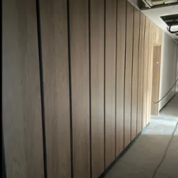 Wykończona ściana z paneli drewnopodobnych z czarnymi wstawkami, widok na sufit z instalacjami w surowym stanie, fragment korytarza z drzwiami.