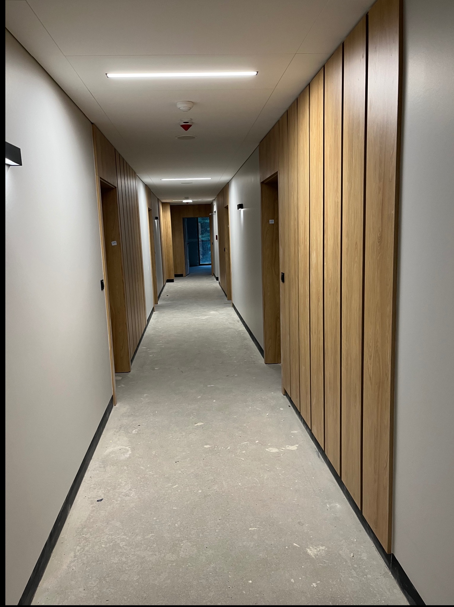 Długi korytarz z drewnianą boazerią i szarymi ścianami. Widoczne drzwi i minimalistyczne oświetlenie. Podłoga surowa, betonowa. Wnętrze w trakcie wykończenia.