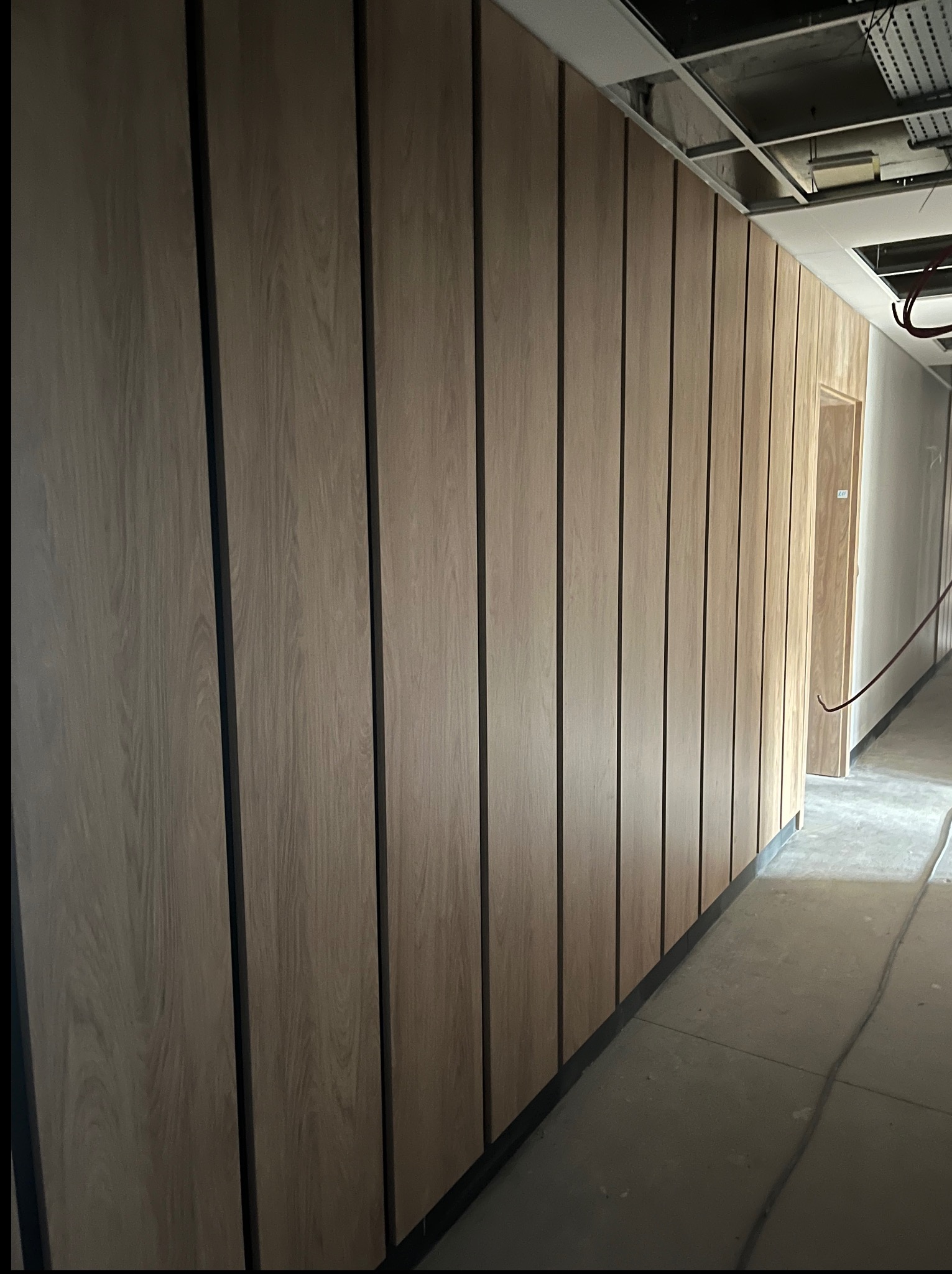 Wykończona ściana z paneli drewnopodobnych z czarnymi wstawkami, widok na sufit z instalacjami w surowym stanie, fragment korytarza z drzwiami.
