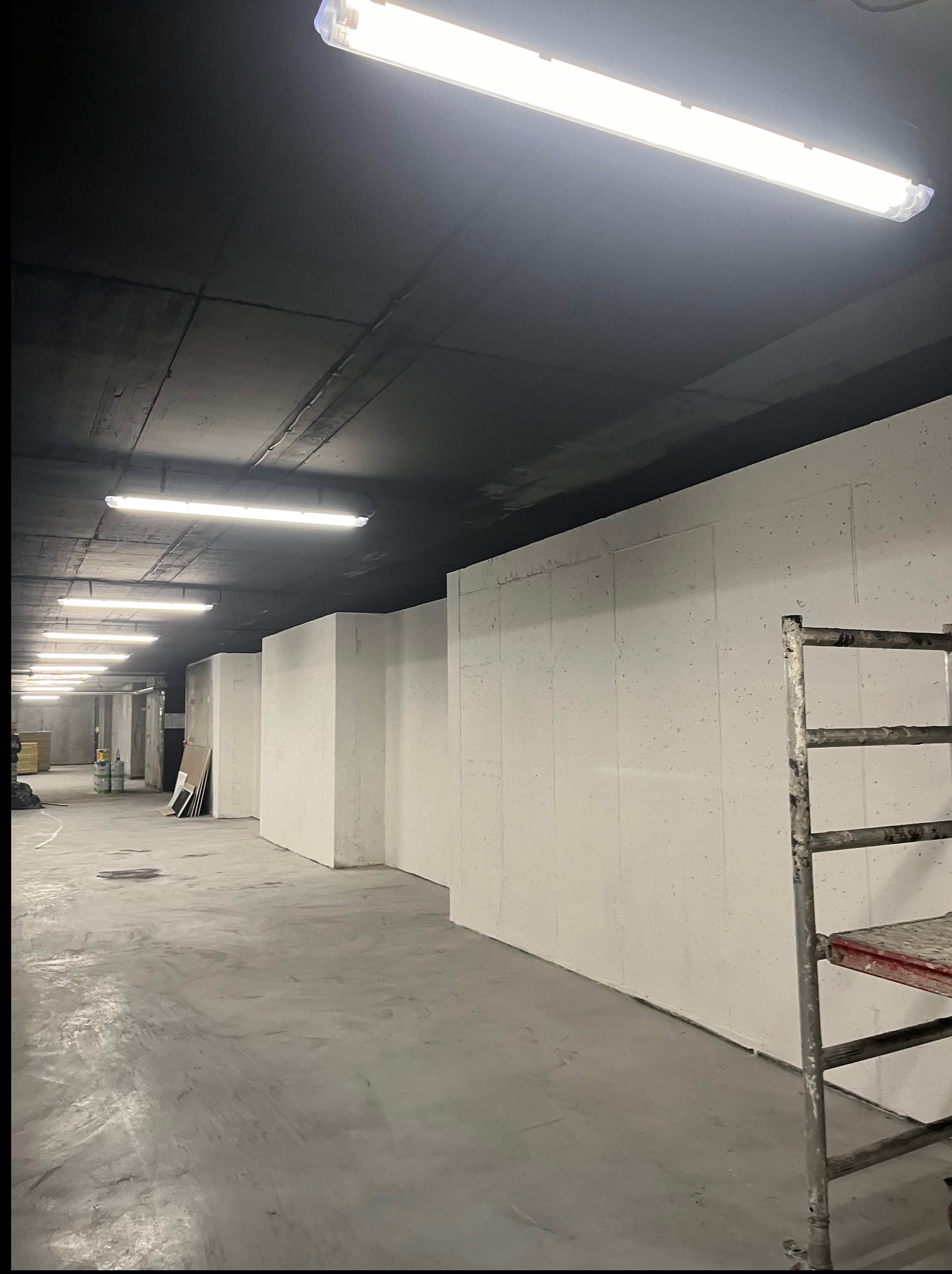 Surowe wnętrze w trakcie prac wykończeniowych, ściany przygotowane do malowania, widoczny betonowy sufit i metalowe rusztowanie. Oświetlenie jarzeniowe.
